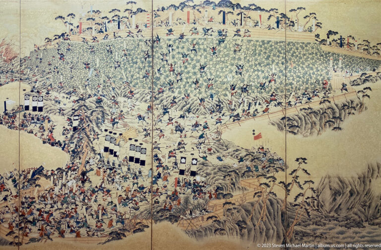 Japanese folding-screen painting with dense soldier formations, mounted figures, standards, tents, and rocky terrain.