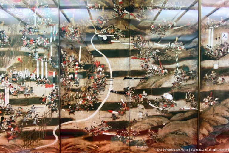 Large Japanese battle screen behind reflective glass, showing warriors, standards, roads, and fortified positions.