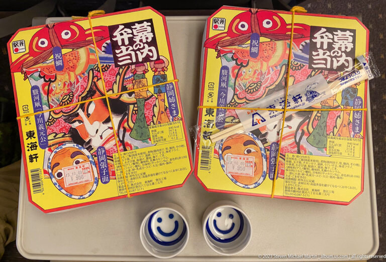 Two colorful Japanese boxed-meal packages beside small smiling-face cups on a tray.