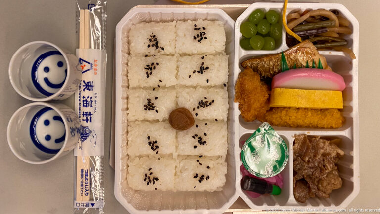 Japanese bento with sesame rice, fish, fried foods, omelet, vegetables, pickles, grapes, meat, cups, and chopsticks.