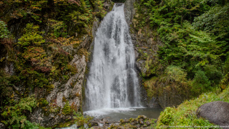 Tall narrow section of Choshi Falls dropping beside mossy rock and green vegetation.