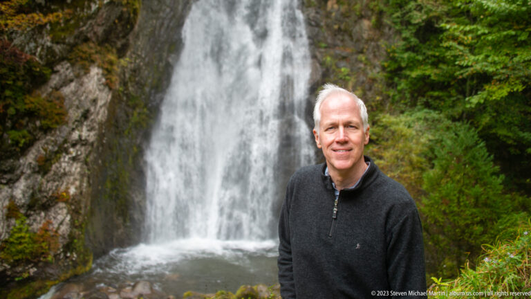 Smiling gray-haired man in a dark sweater standing before a blurred waterfall.