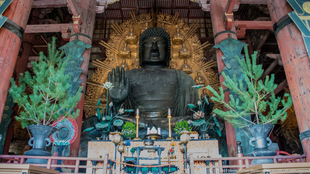 Frontal view of Todai-ji’s bronze Daibutsu above an altar with flowers, candles, vessels, and greenery.