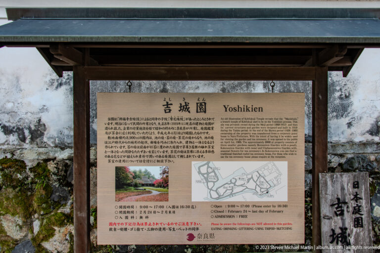 Yoshikien garden information panel beneath a timber shelter beside a stone wall.