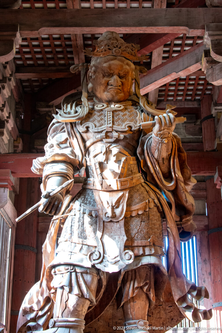 Monumental wooden guardian deity in armor standing beneath the timber roof of the Great Buddha Hall.