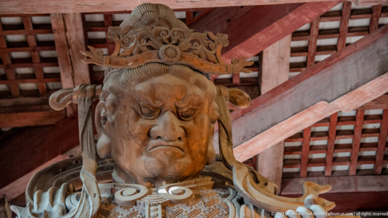 Close view of a fierce carved wooden guardian face with elaborate headdress beneath red timber beams.