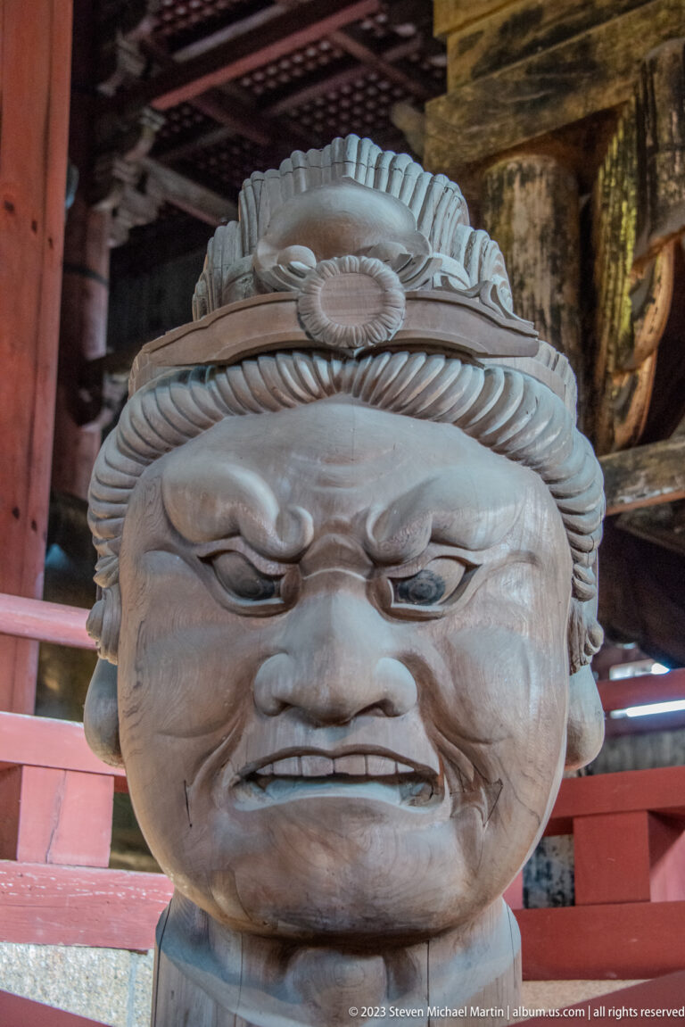 Frontal close-up of a monumental wooden guardian head with bulging eyes and layered headdress.