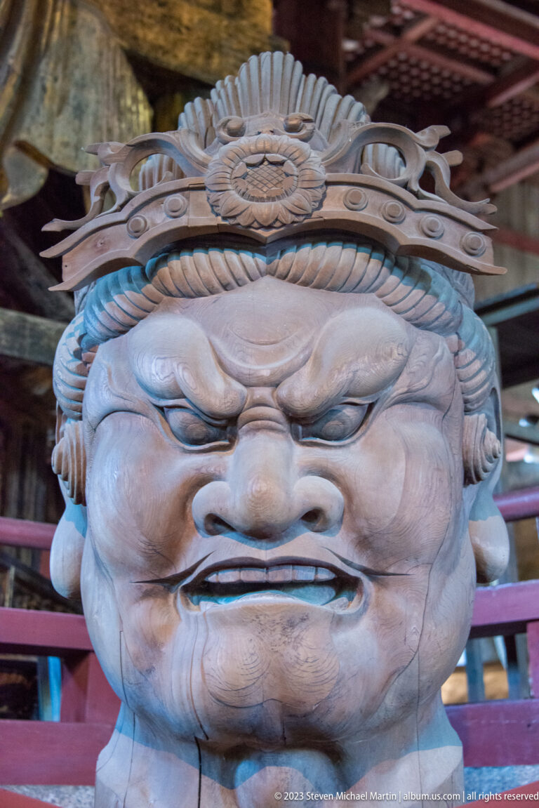 Frontal close-up of a fierce wooden Buddhist guardian with bulging eyes and braided headdress.