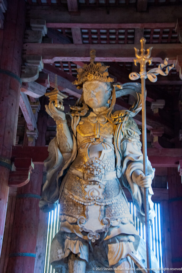 Monumental armored Buddhist guardian holding a tall staff and a small pagoda-like object.