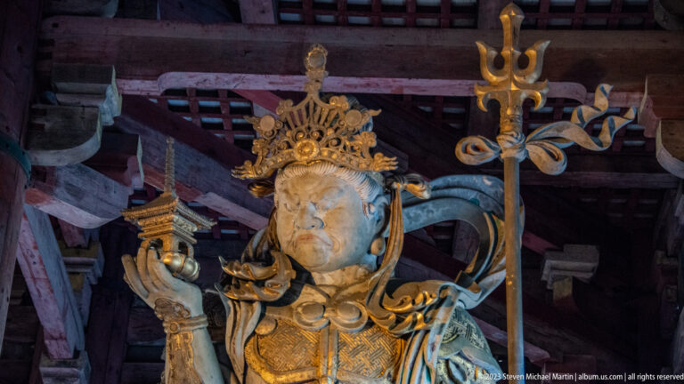 Close view of crowned armored guardian holding a trident-like staff and small pagoda.