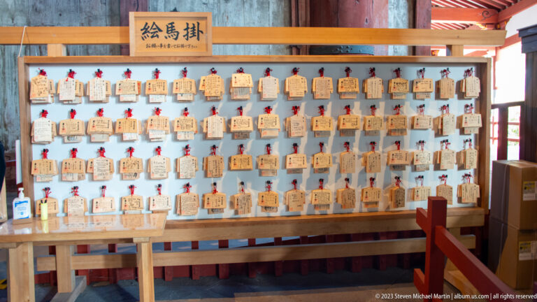 Rows of wooden ema votive plaques with handwritten messages hanging from red cords.