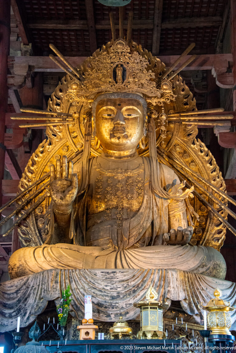 Frontal view of a monumental gilded seated attendant with raised hand and radiating aureole.