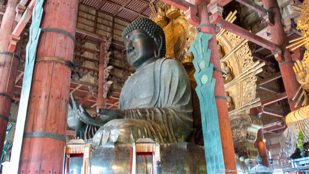 Oblique profile of Todai-ji’s bronze Daibutsu between red columns and a turquoise banner.