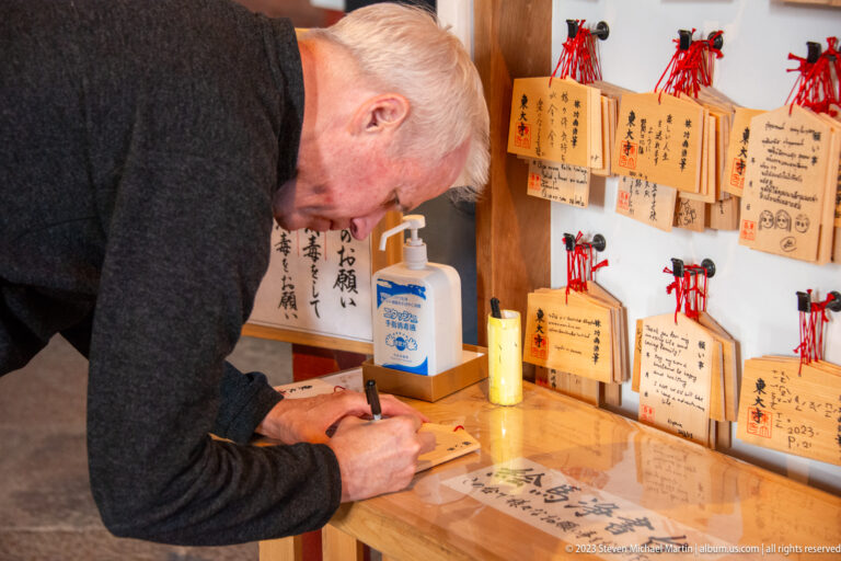 Visitor writing on a wooden ema plaque beside a wall of completed votive tablets.