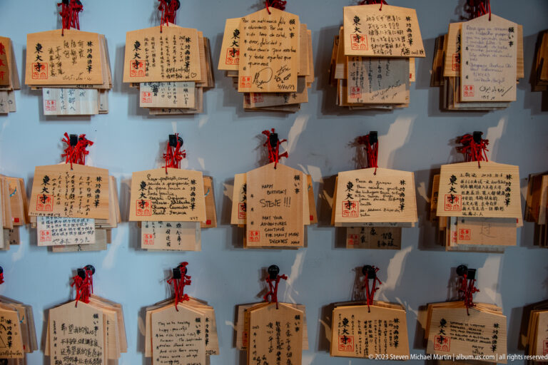Wall filled with rows of wooden ema plaques carrying multilingual handwritten wishes and drawings.