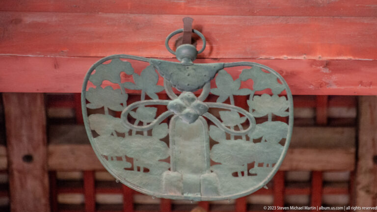 Openwork metal pendant with paired deer and curling plant motifs on a red timber beam.
