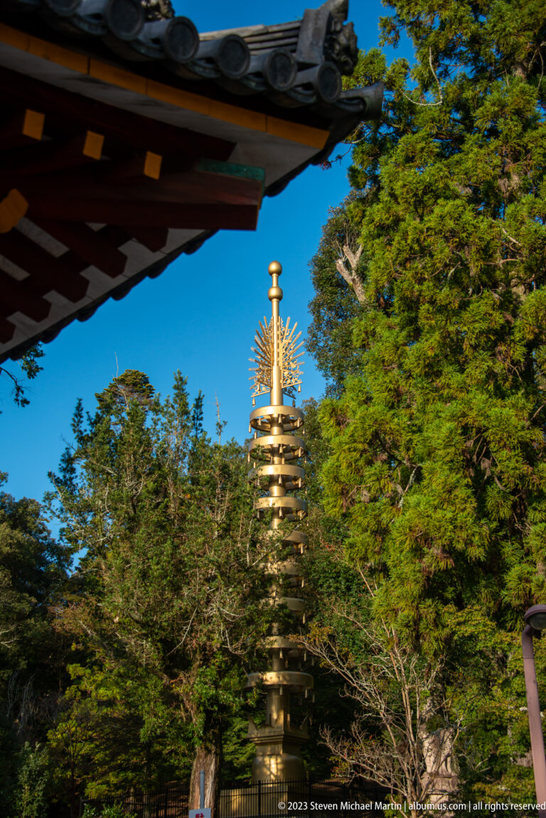 Tall bronze pagoda-form monument with stacked tiers rising among trees beneath a blue sky.