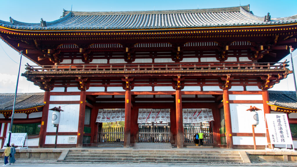 Large vermilion two-story temple gate with white panels, tiled roof, and open central bays.