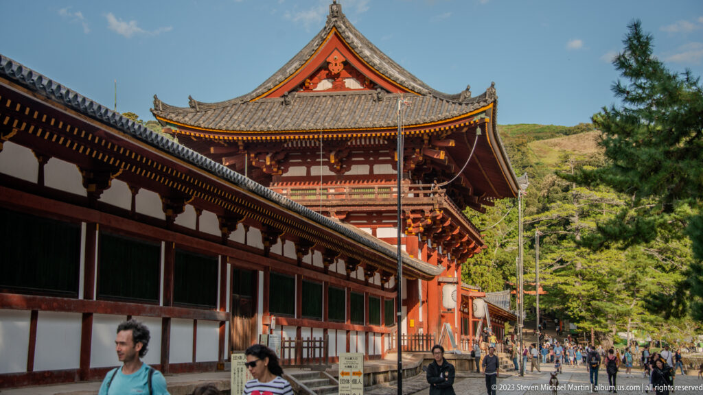 Vermilion timber temple hall rising behind a long covered corridor with visitors in the foreground.