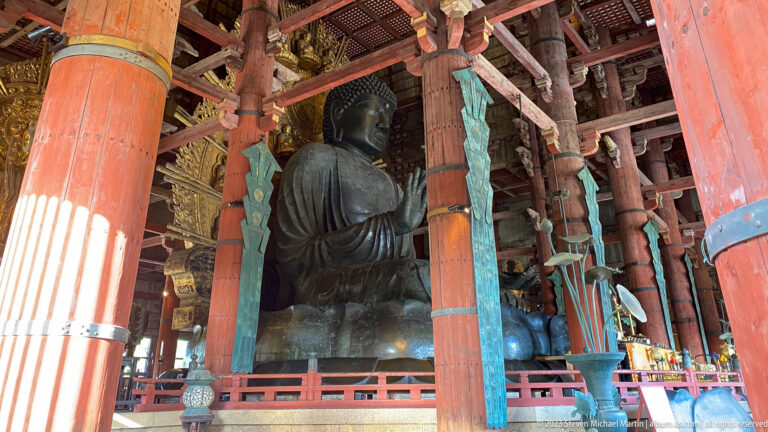 Oblique view of Todai-ji’s bronze Daibutsu framed by red columns, beams, and turquoise hangings.