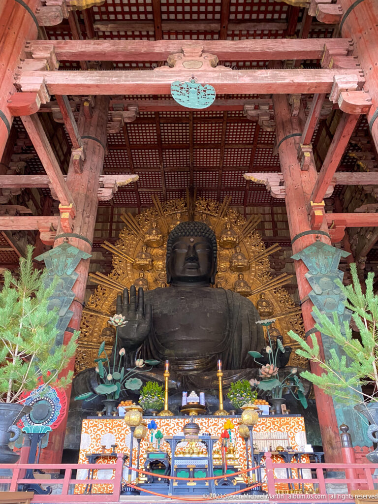 Frontal view of Todai-ji’s Daibutsu above an altar with candles, flowers, vessels, textiles, and greenery.