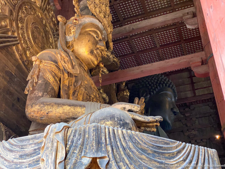Gilded seated bodhisattva attendant in the foreground with the dark bronze Daibutsu visible behind.