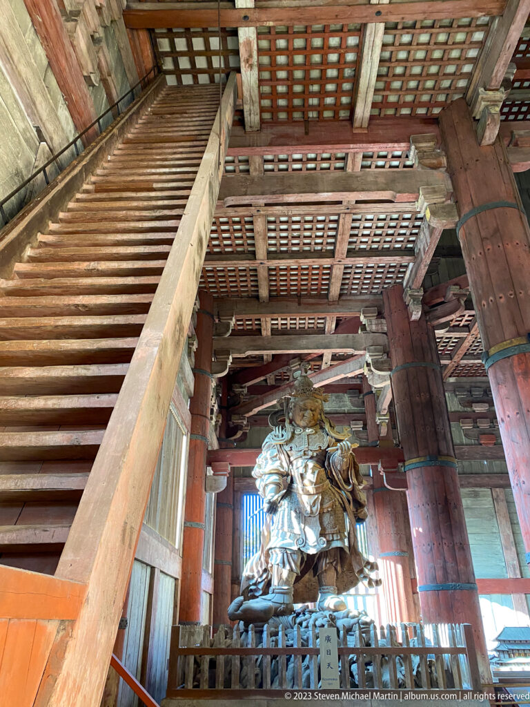 Steep timber staircase rising beside a monumental wooden guardian beneath the hall’s lattice ceiling.