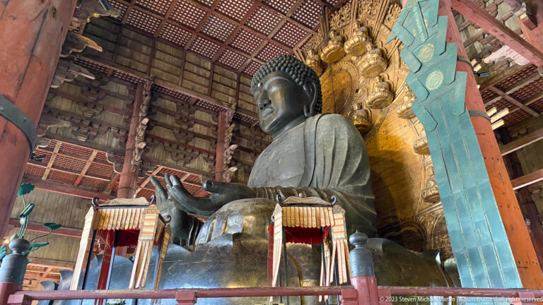 Low oblique view of Todai-ji’s bronze Daibutsu with open palm, gilded halo, and turquoise hanging.