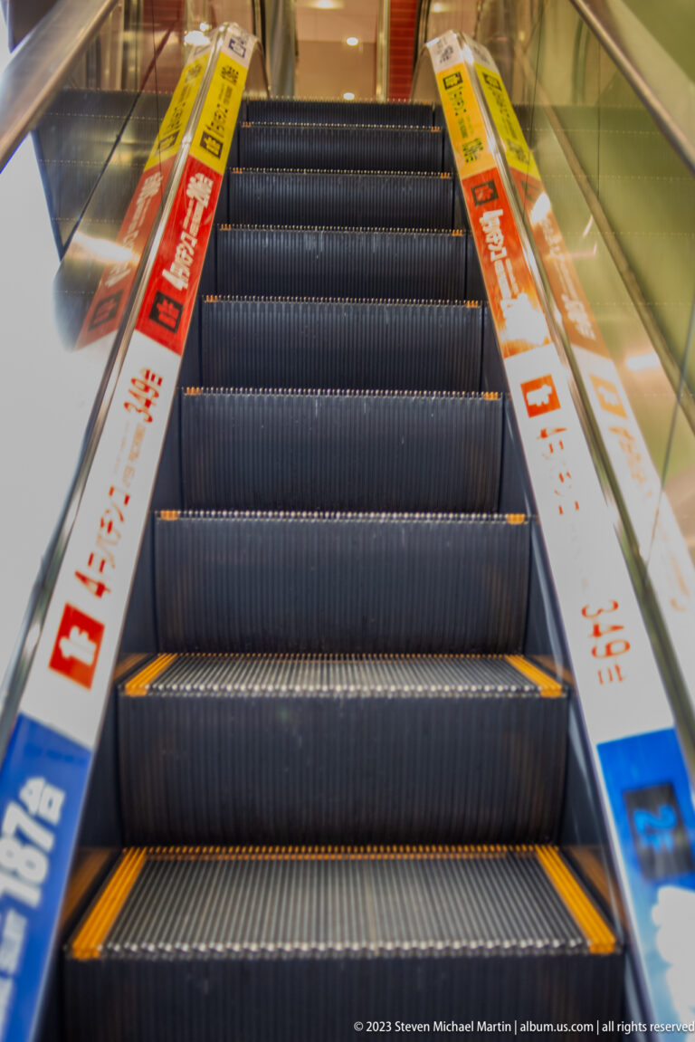 Upward indoor escalator bordered by Japanese advertising strips on both sides.
