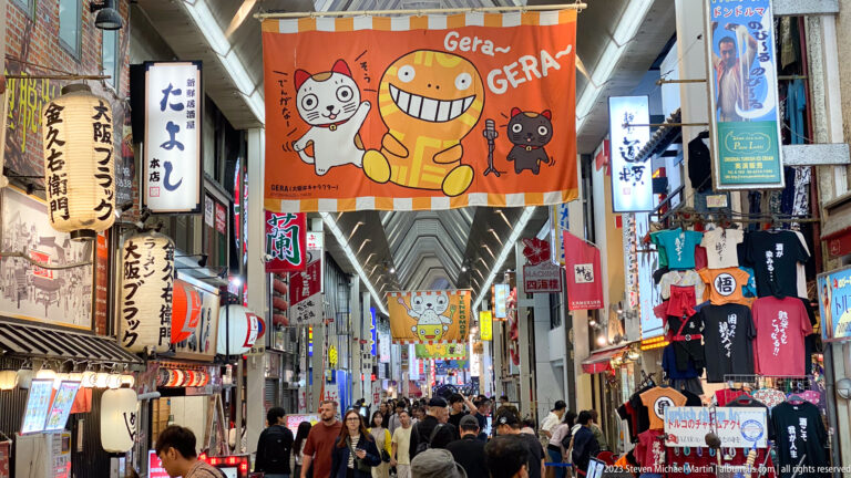 Busy covered Osaka shopping arcade beneath a cartoon cat banner and hanging signs.
