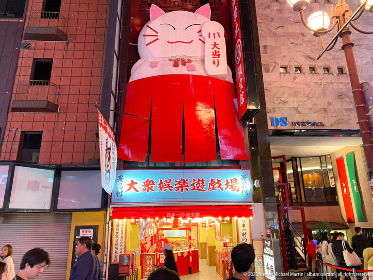 Large white and red beckoning cat figure above a brightly lit Osaka arcade entrance.