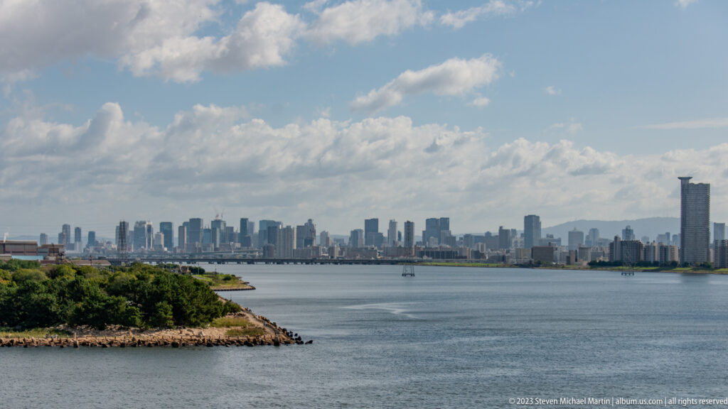 Osaka skyline across broad water beneath a blue sky with scattered clouds.