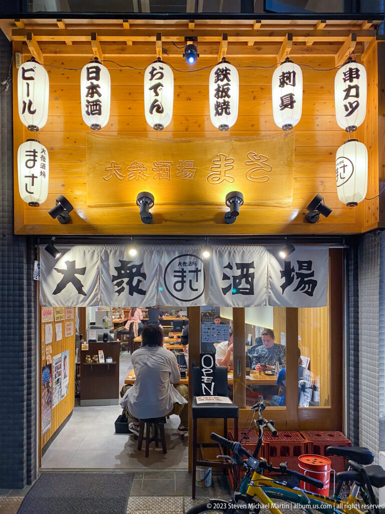 Open Osaka izakaya with lanterns, noren curtains, and patrons at a counter.
