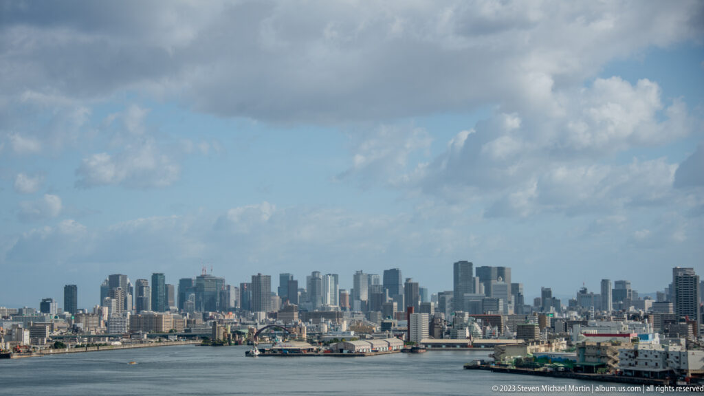 Wide Osaka harbor panorama with skyline, cranes, bridges, and heavy clouds.