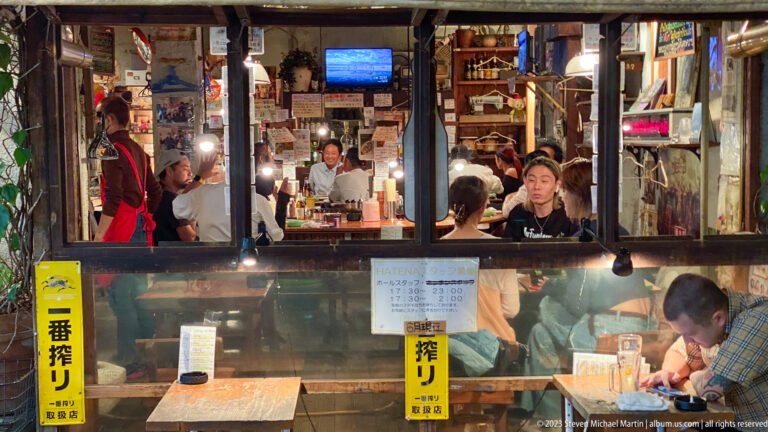 Busy open-front Osaka izakaya with diners, staff, posters, and bottles visible inside.