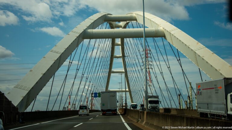 Vehicles crossing beneath the white steel arches of a large Osaka bridge.
