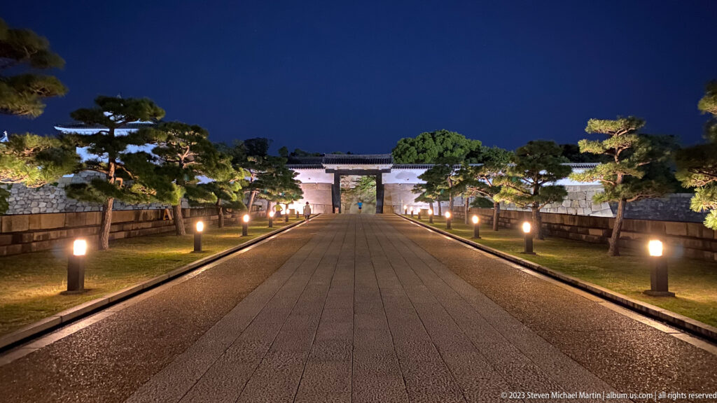 Symmetrical lit path leading toward a gate in the Osaka Castle grounds at night.