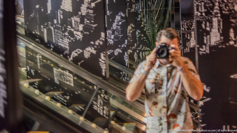 Blurred photographer reflected in glass before a large black-and-white city map graphic.