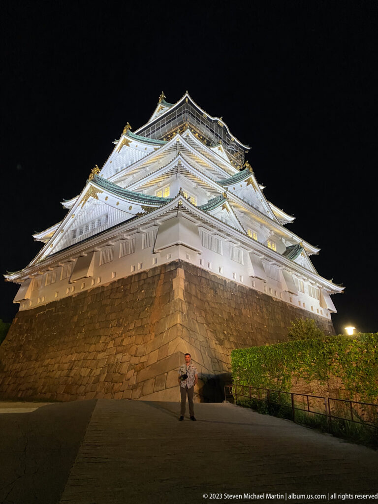 Low-angle view of illuminated Osaka Castle above a massive stone base with one visitor.