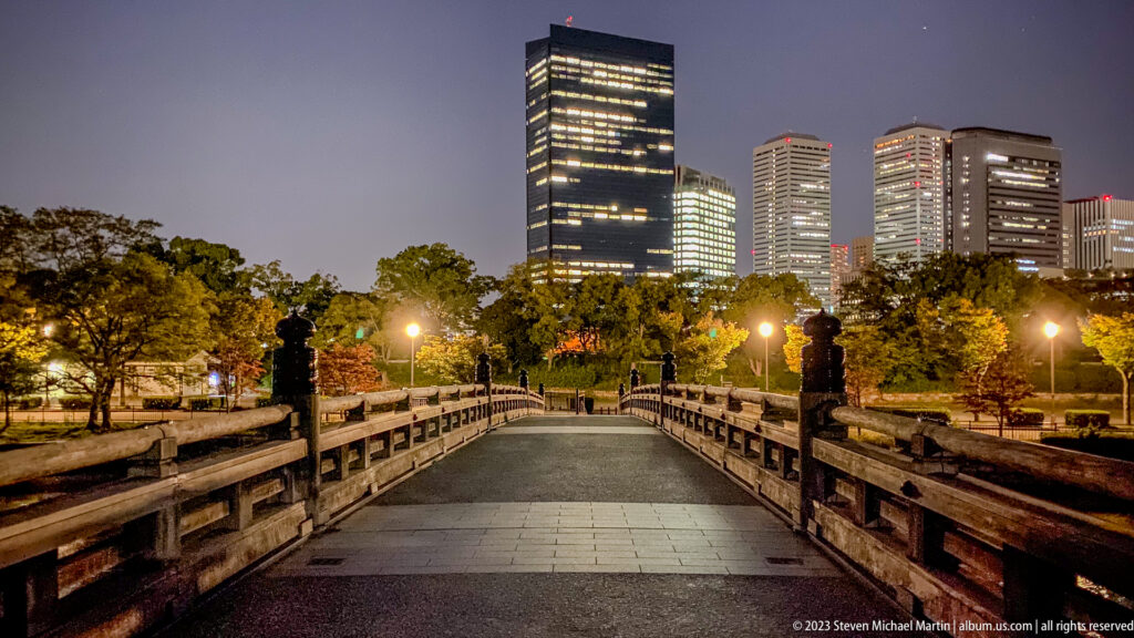 Stone bridge in Osaka Castle Park leading toward illuminated modern towers at night.