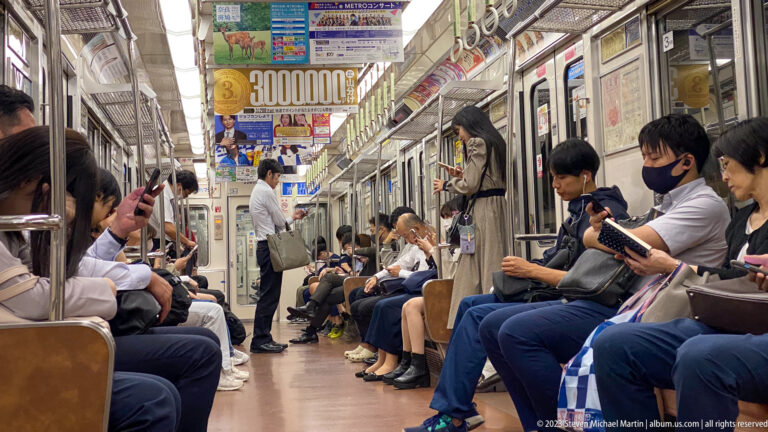 Passengers seated and standing inside a crowded Osaka commuter train beneath hanging straps and advertisements.