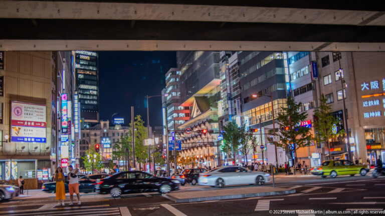 Cars and pedestrians along a brightly lit commercial avenue in central Osaka at night.