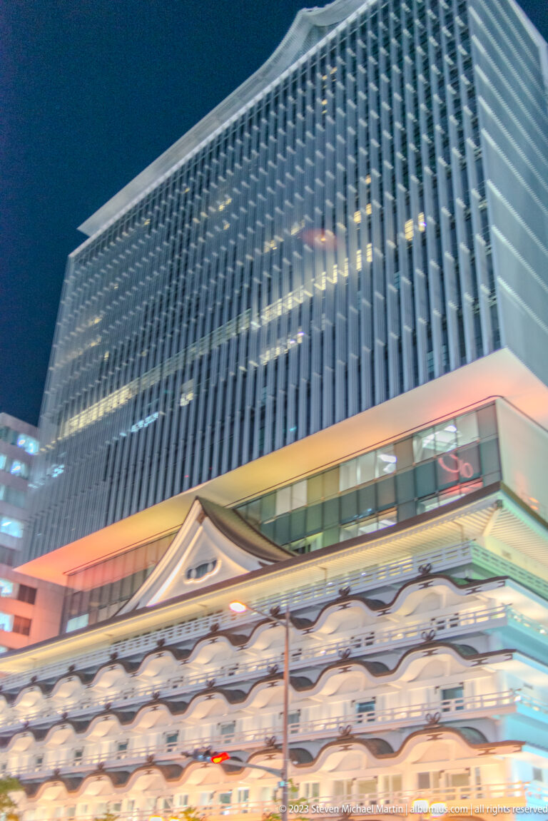 Modern gridded building above a lower traditional-style roofline in Osaka at night.