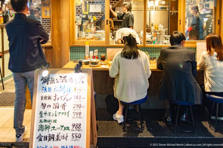 Diners at a small Osaka counter restaurant beside a handwritten seafood menu.