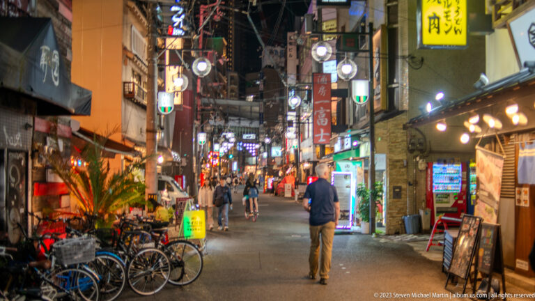 Narrow Osaka nightlife alley with signs, bicycles, vending machines, and one pedestrian.