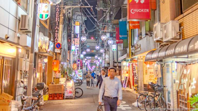 Bright Osaka backstreet lined with small restaurants, signs, bicycles, and pedestrians.