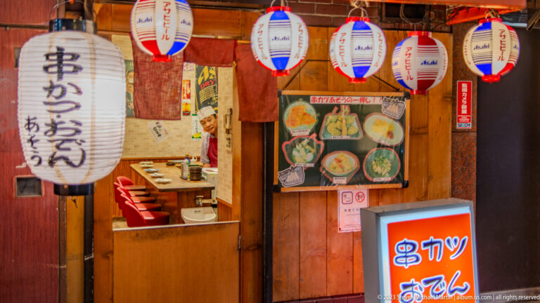 Small Osaka restaurant entrance with lanterns, menu photos, and signs for kushikatsu and oden.