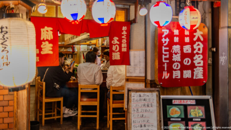 Diners seated in a compact Osaka izakaya beneath red curtains and paper lanterns.