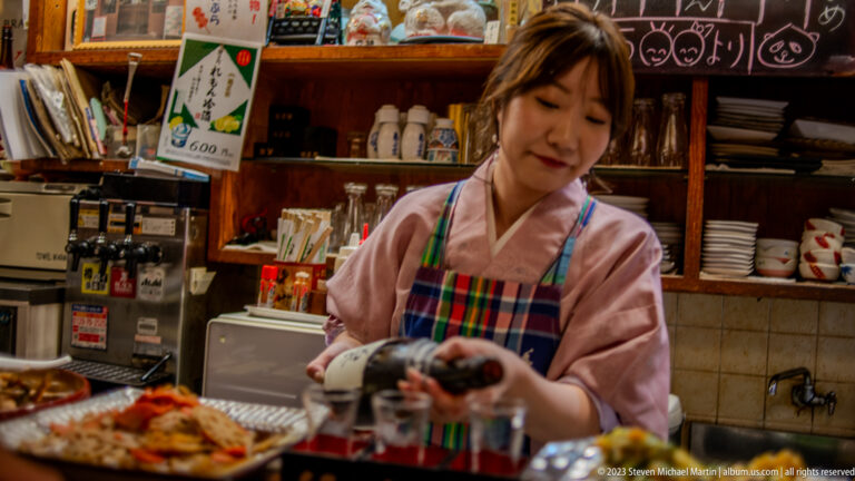 Restaurant server in a kimono-style uniform holding a dark ceramic bottle behind the counter.