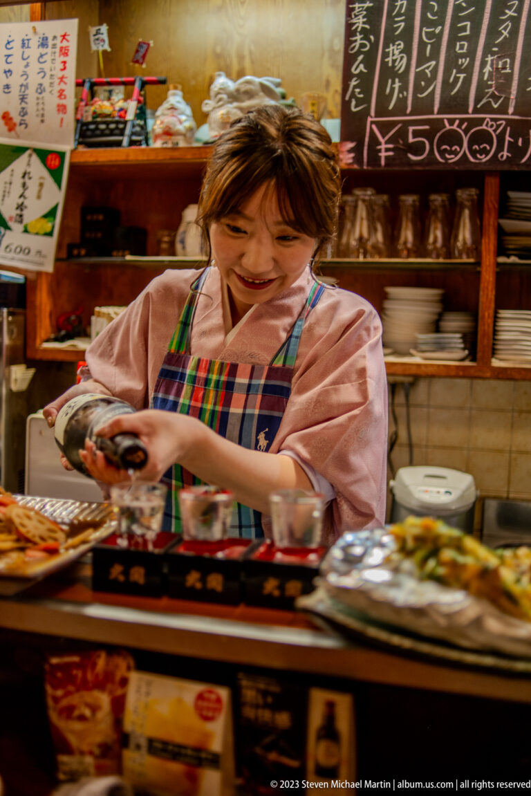 Server in a kimono-style uniform preparing small dishes behind an Osaka restaurant counter.